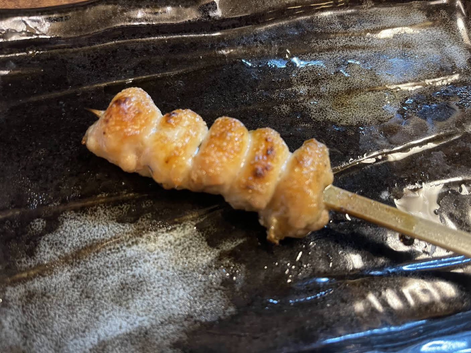 A chicken skewer with evenly sized pieces on a black plate at Yakitori Senkichi