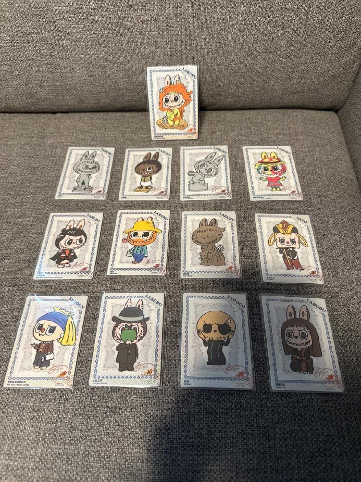 Thirteen sleeved LABUBU art cards laid out on a grey sofa
