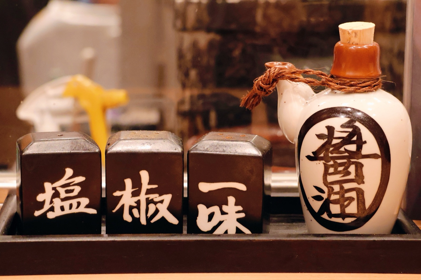 Black containers labelled 塩, 椒 and 一味 on a wooden stand, next to a white flask labelled 醤油