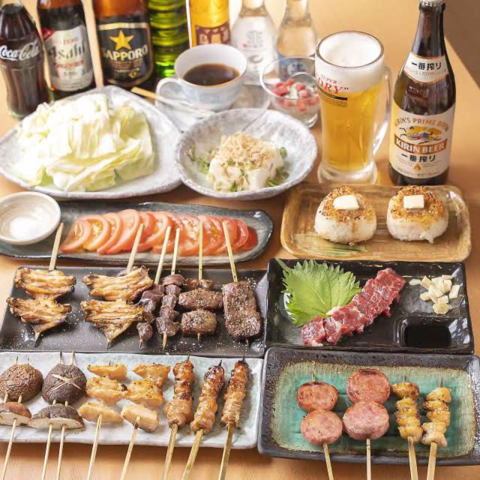 テーブルに並んだ串焼き、馬刺し、焼きおにぎり、キャベツ、トマトなどの料理と飲み物