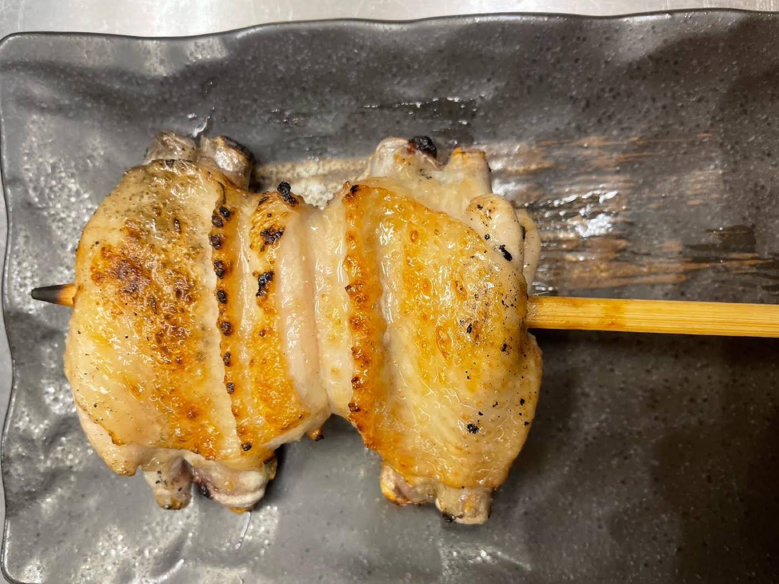 A chicken wing on a bamboo skewer, the skin browned from the grill, on a grey rectangular plate
