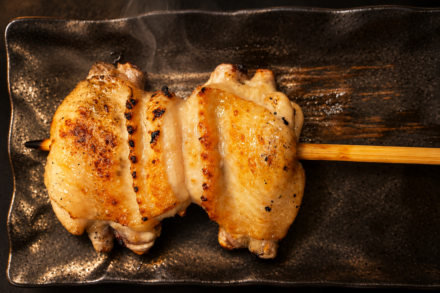 暗い角皿の上で、皮目に焼き色がついた鶏の手羽の串焼き