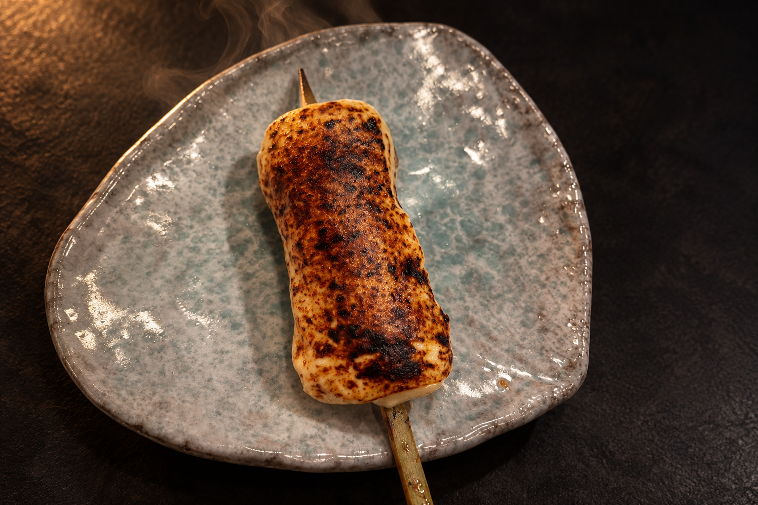 A seared, lightly charred skewer on a pale speckled plate, with a little steam rising