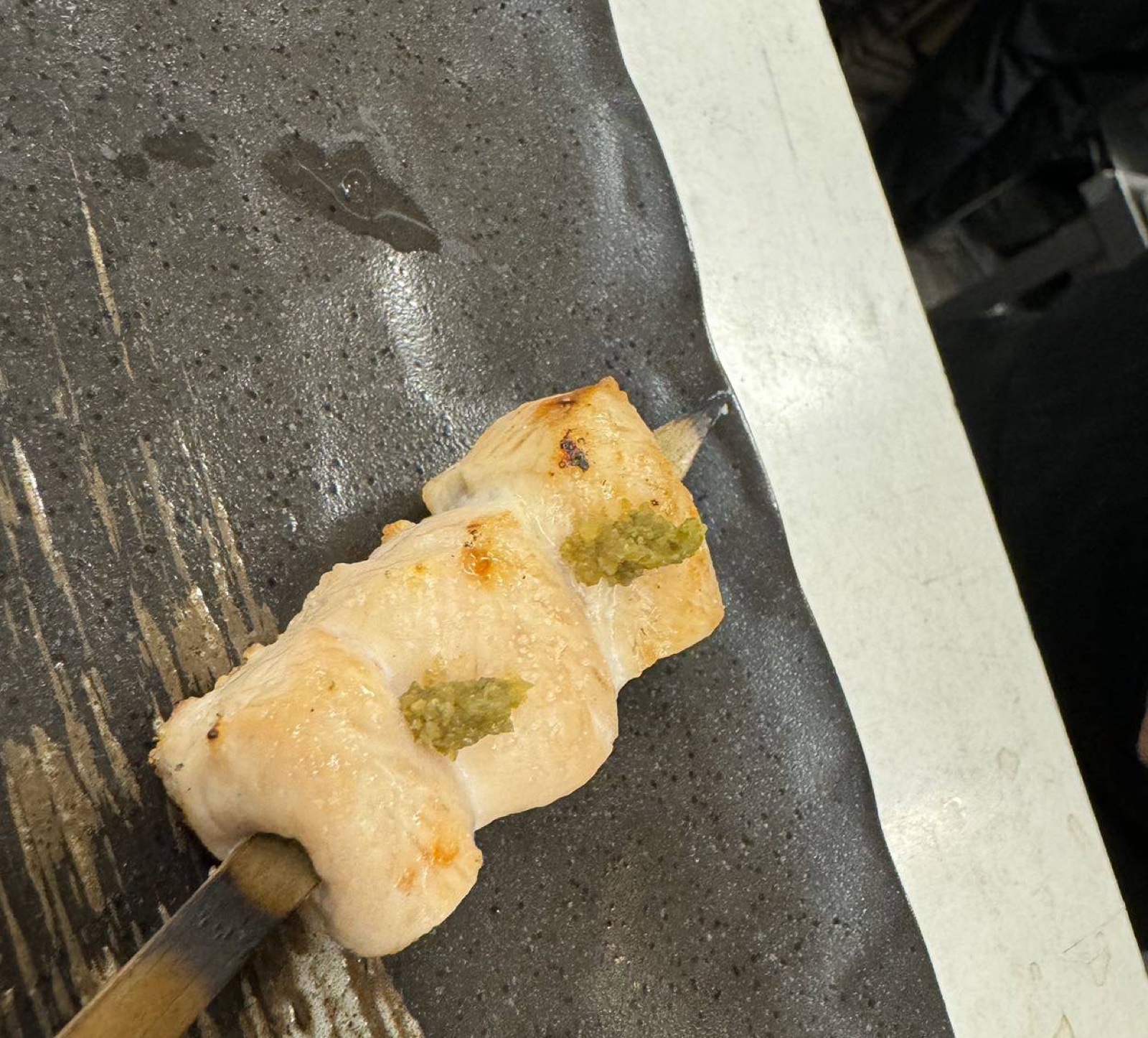 A white chicken skewer with two dabs of wasabi on a black plate