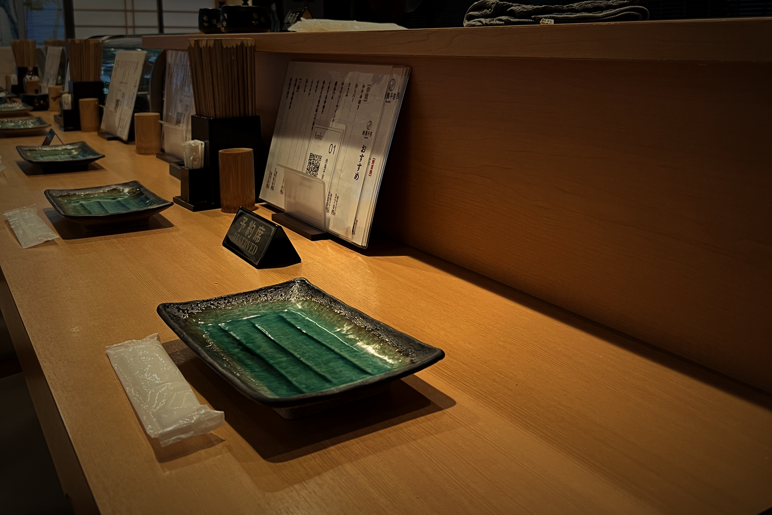 The wooden counter at Yakitori Senkichi in warm low light, with green glazed plates, a reserved sign and a menu stand