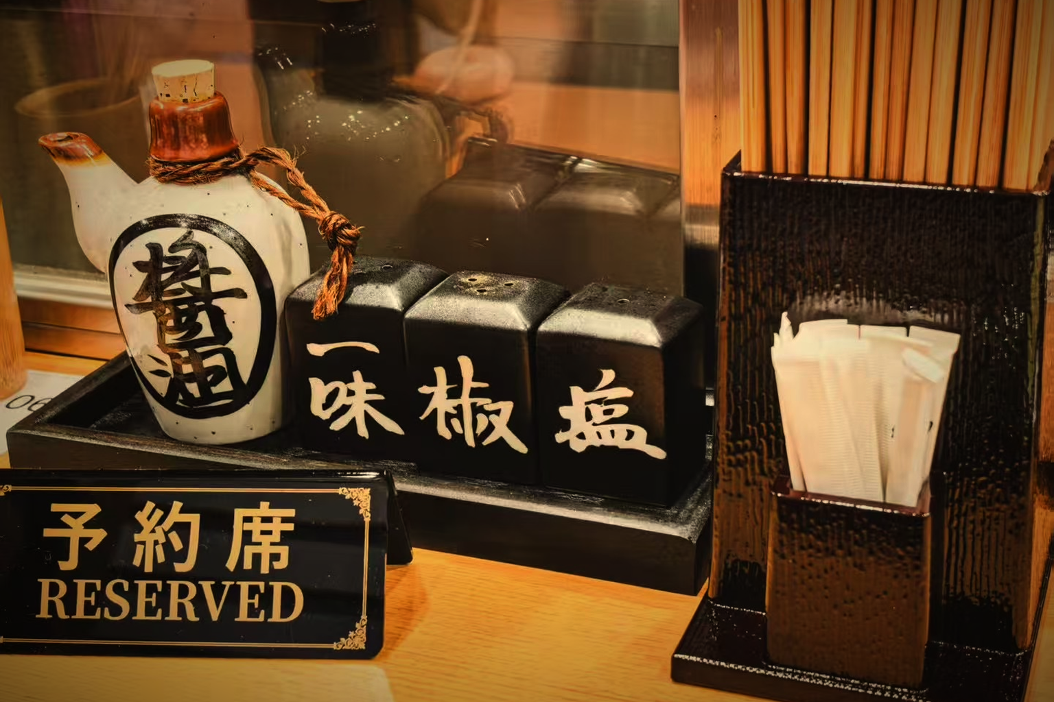 A soy sauce pot and three shakers labelled ichimi, sansho and salt on a dark tray at Yakitori Senkichi, with a reserved sign and chopsticks in warm low light
