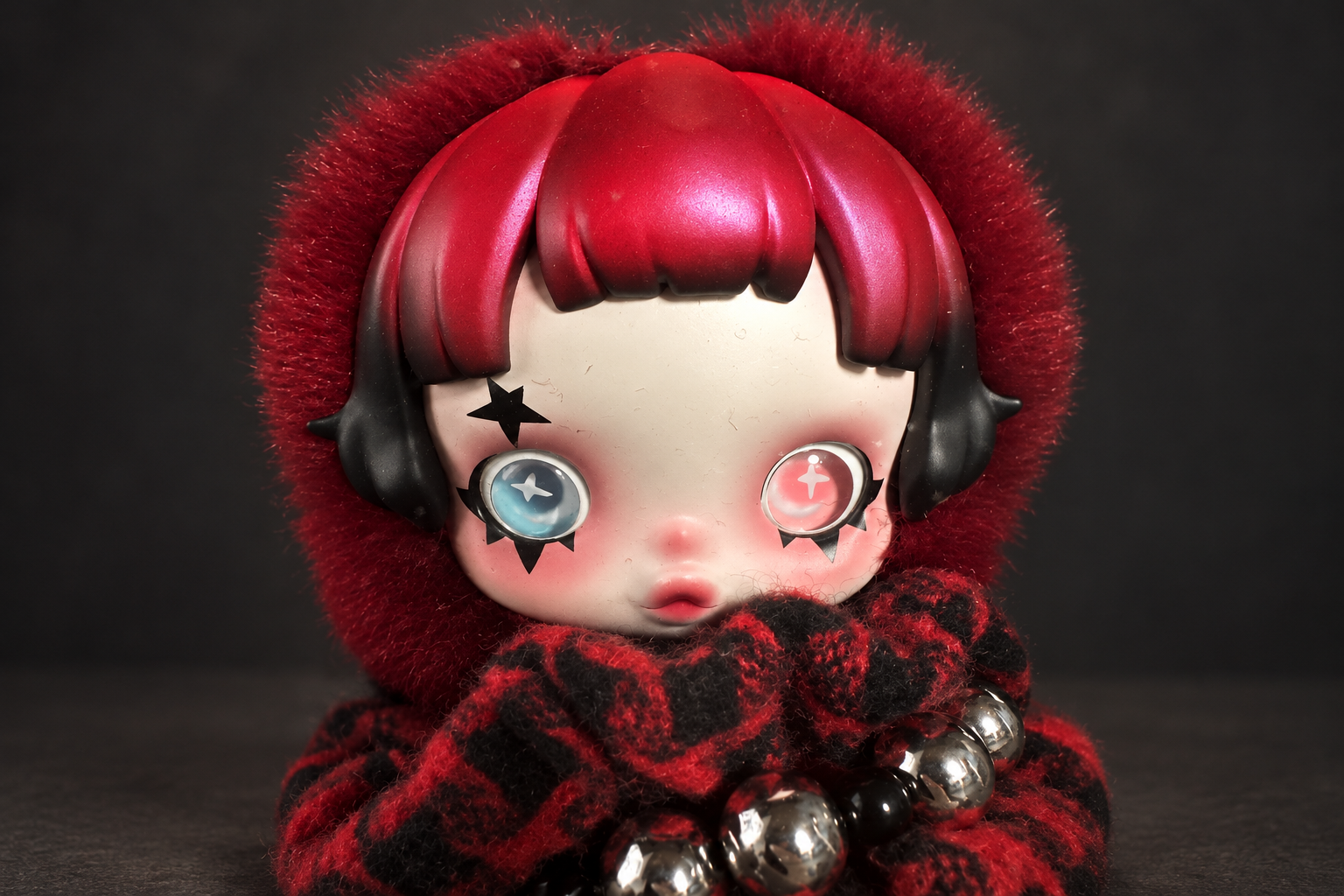 Close-up of the SKULLPANDA 6kHz plush's face, with red bobbed hair, blue and pink eyes, a red fur hood and a red-and-black check scarf with silver beads, on a dark grey background