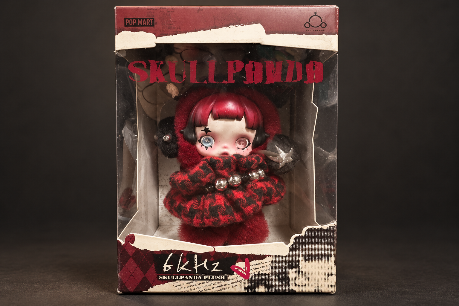 深いグレーの背景に置いた、透明窓つきのSKULLPANDA 6kHzぬいぐるみの箱。赤い髪と赤黒チェックの衣装が見える