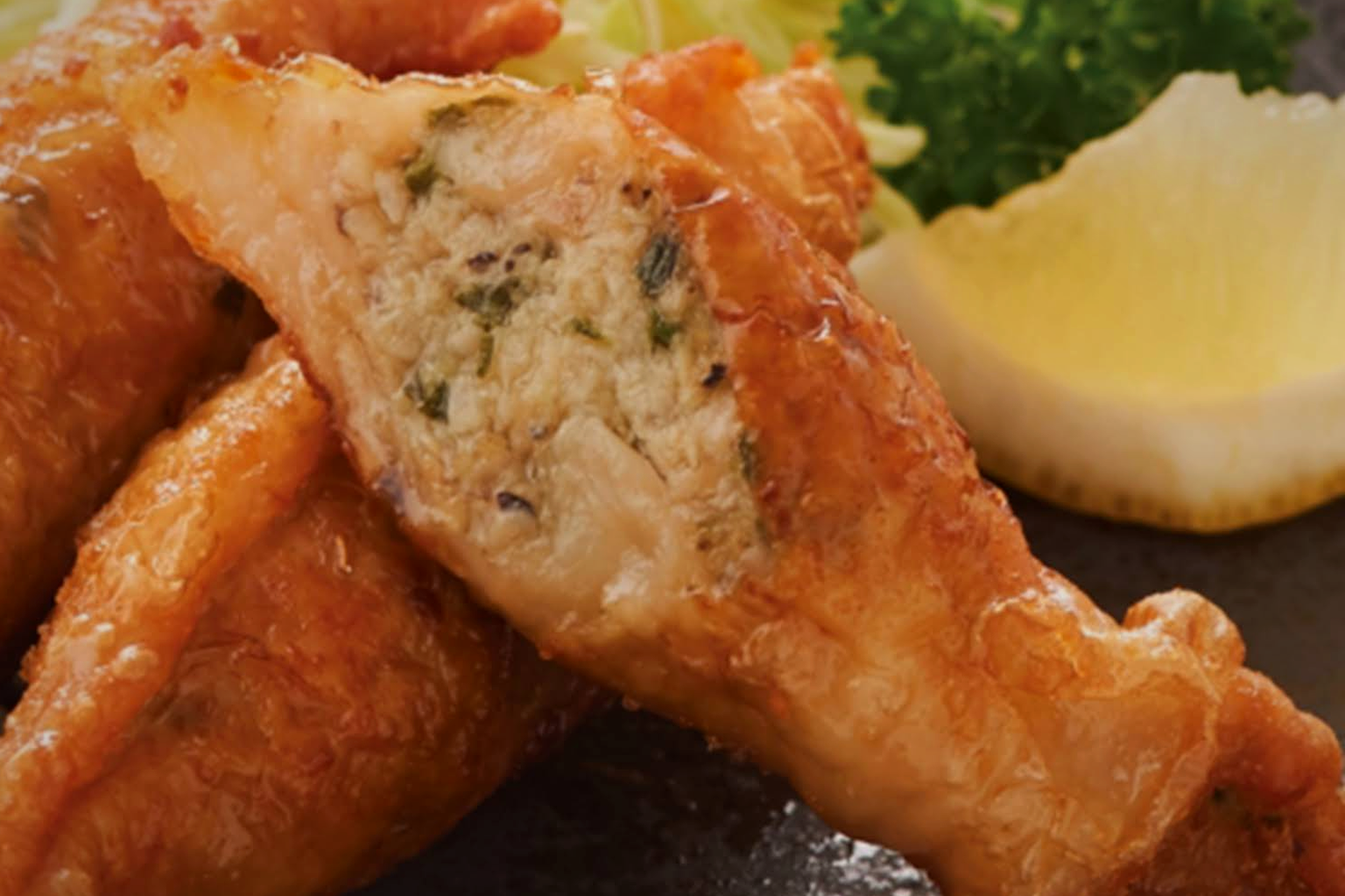 Close-up of a halved tebagyoza showing the minced filling flecked with green, next to golden fried wings and a lemon wedge