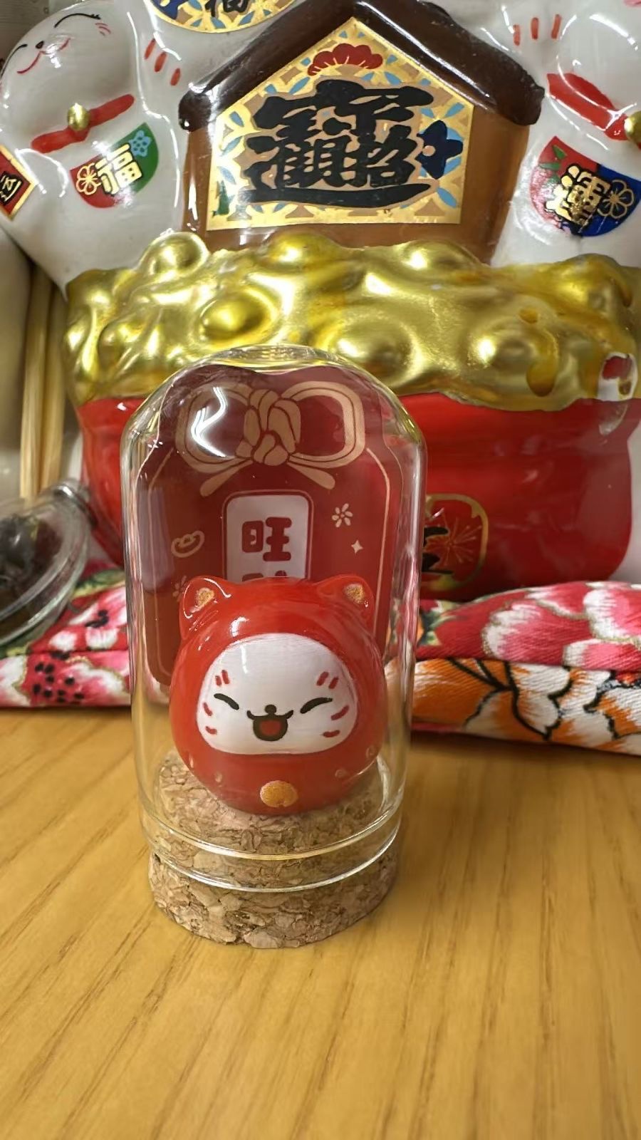 The red daruma cat in its glass dome on a cork base, with a red card showing 旺 behind it; the large cat figure in the back is a photo prop