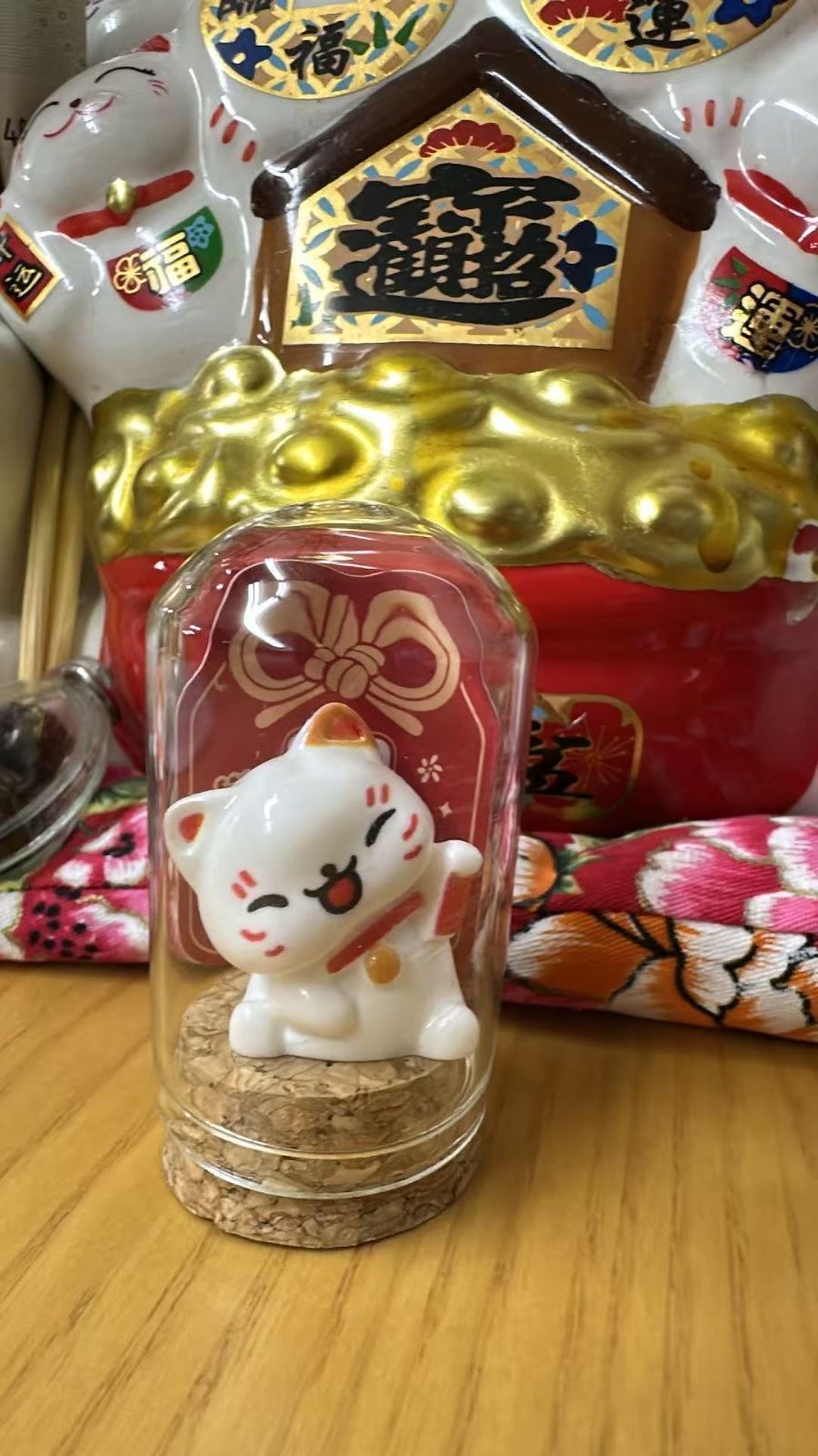 The white beckoning cat in its glass dome on a cork base, raising the paw on the right as you face it and holding a small red item