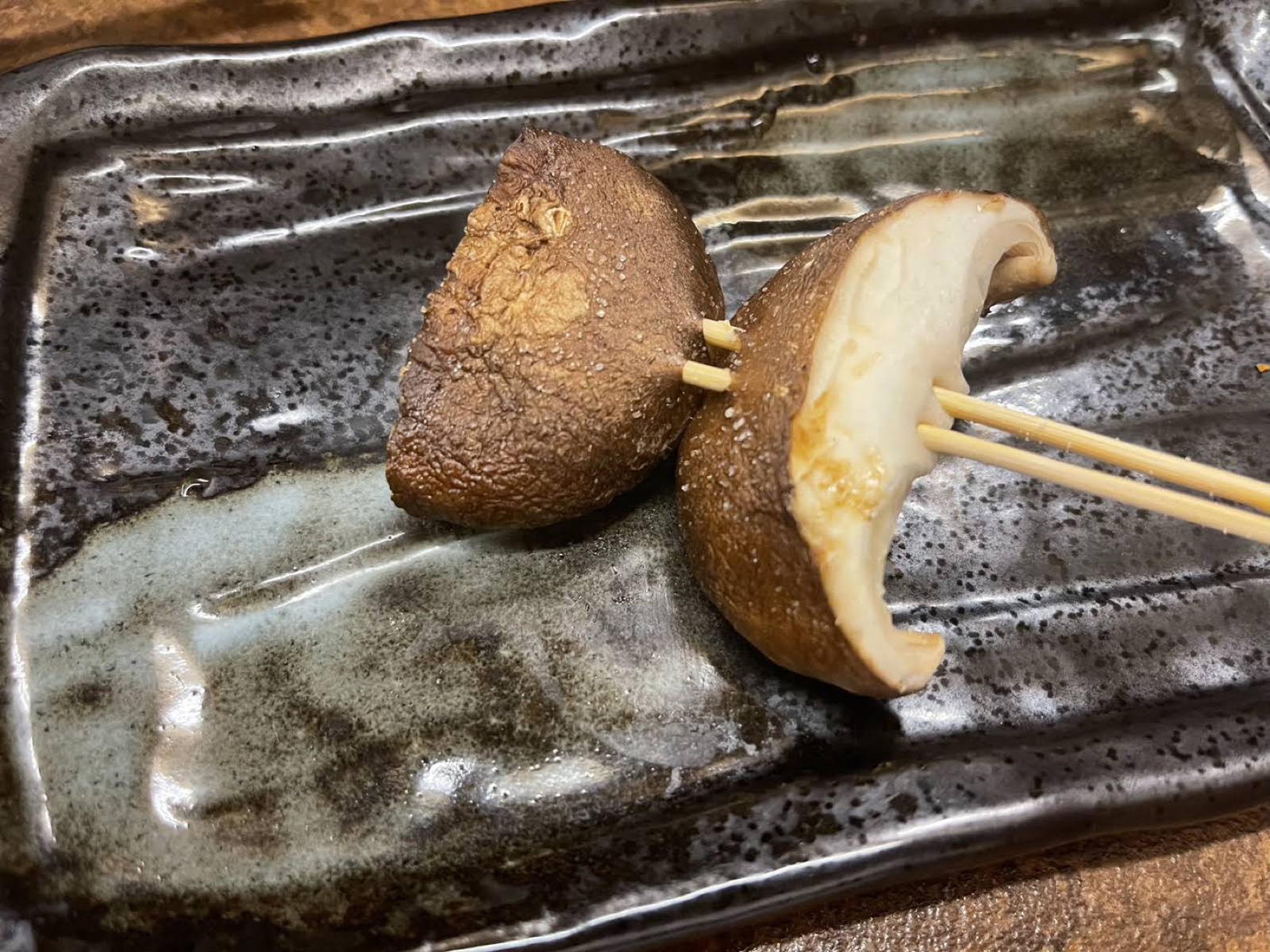 A halved shiitake cap on two skewers on a dark glazed plate, salted, with the cut face visible (unedited photo)
