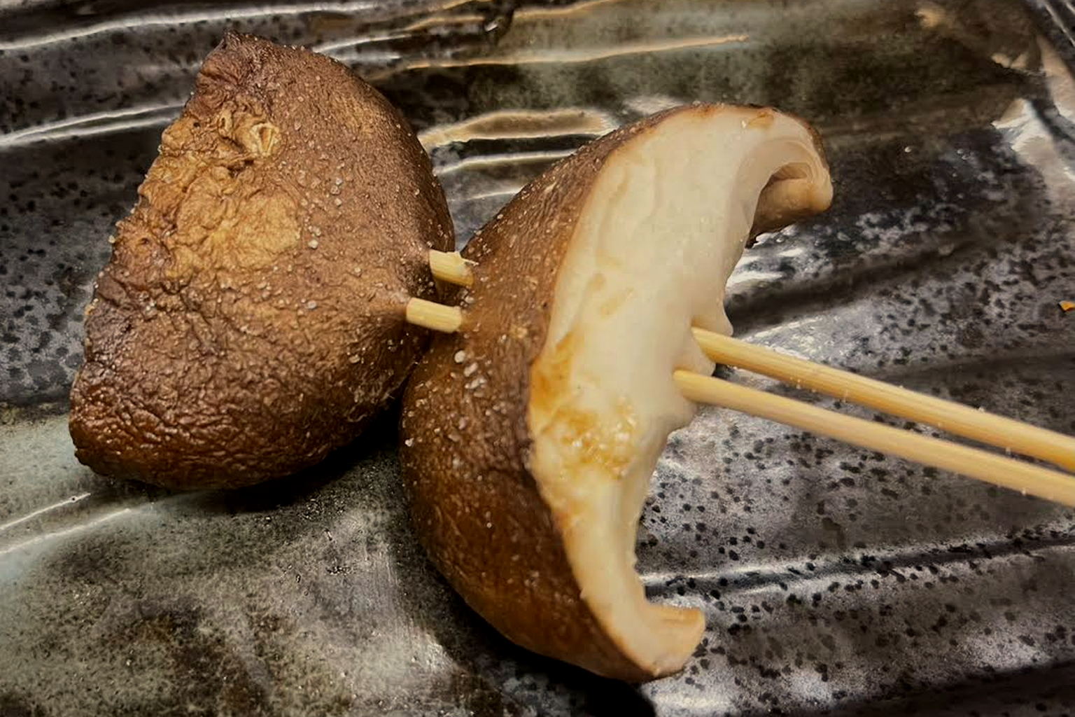 Close-up of a halved shiitake cap on a double skewer, salt crystals on the brown cap and the cut face cooked through to white