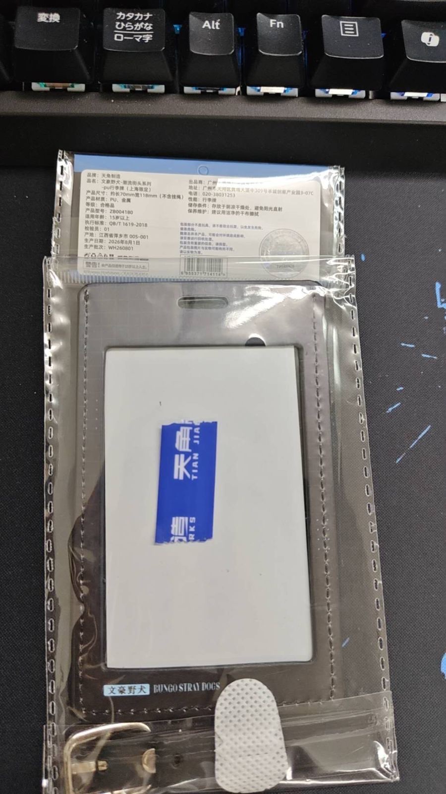 文豪ストレイドッグスのPUラゲッジタグを袋の裏側から撮った写真。上部のラベルに品牌、品名、产品尺寸、产品材质、适用年龄、执行标准、产地、生产日期などが中国語で書かれている