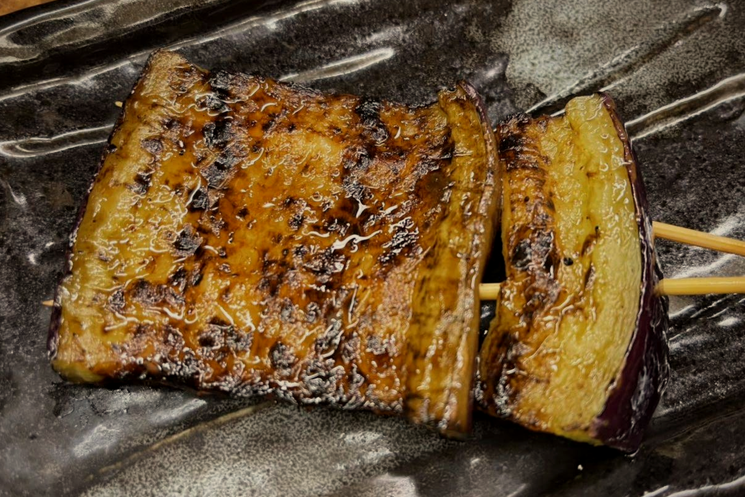 Close-up of two large pieces of eggplant on a double skewer, glossy with tare and charred in places, purple skin visible at the edge, on a dark glazed plate
