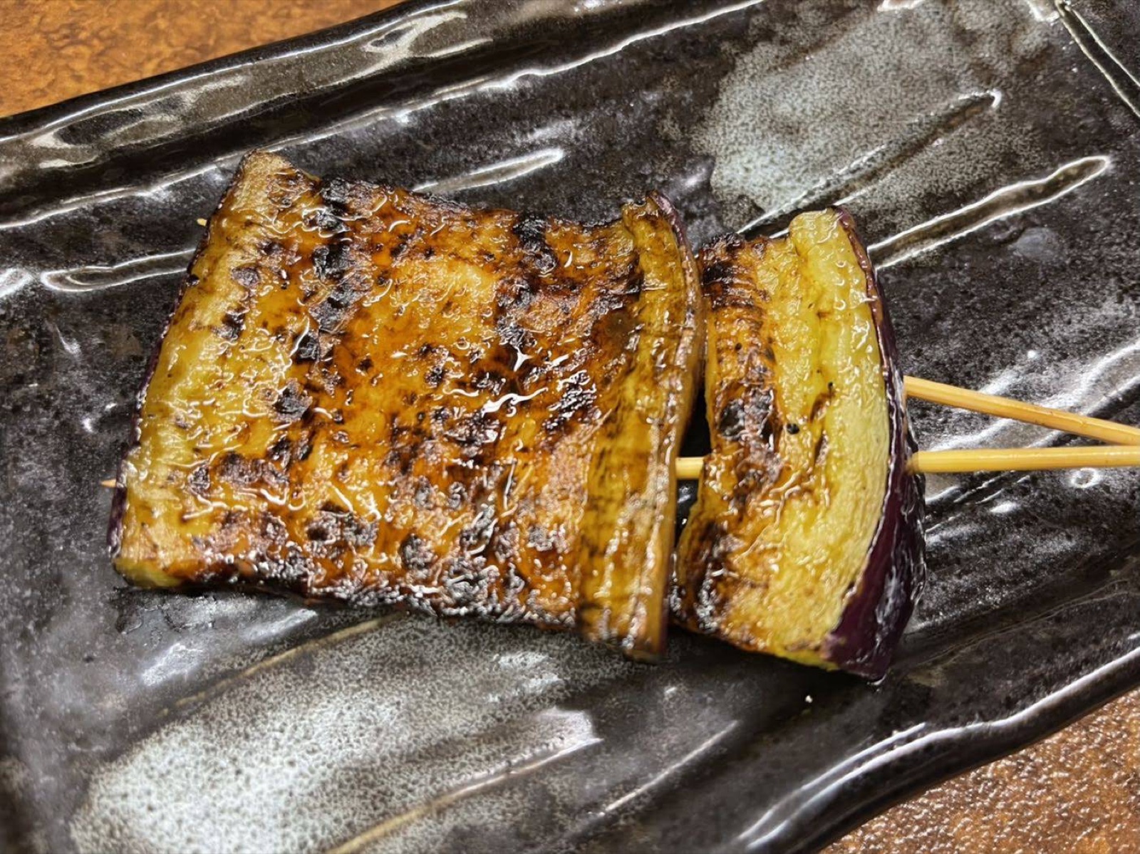 A tare-glazed eggplant skewer held on two skewers on a dark glazed plate (unedited photo)