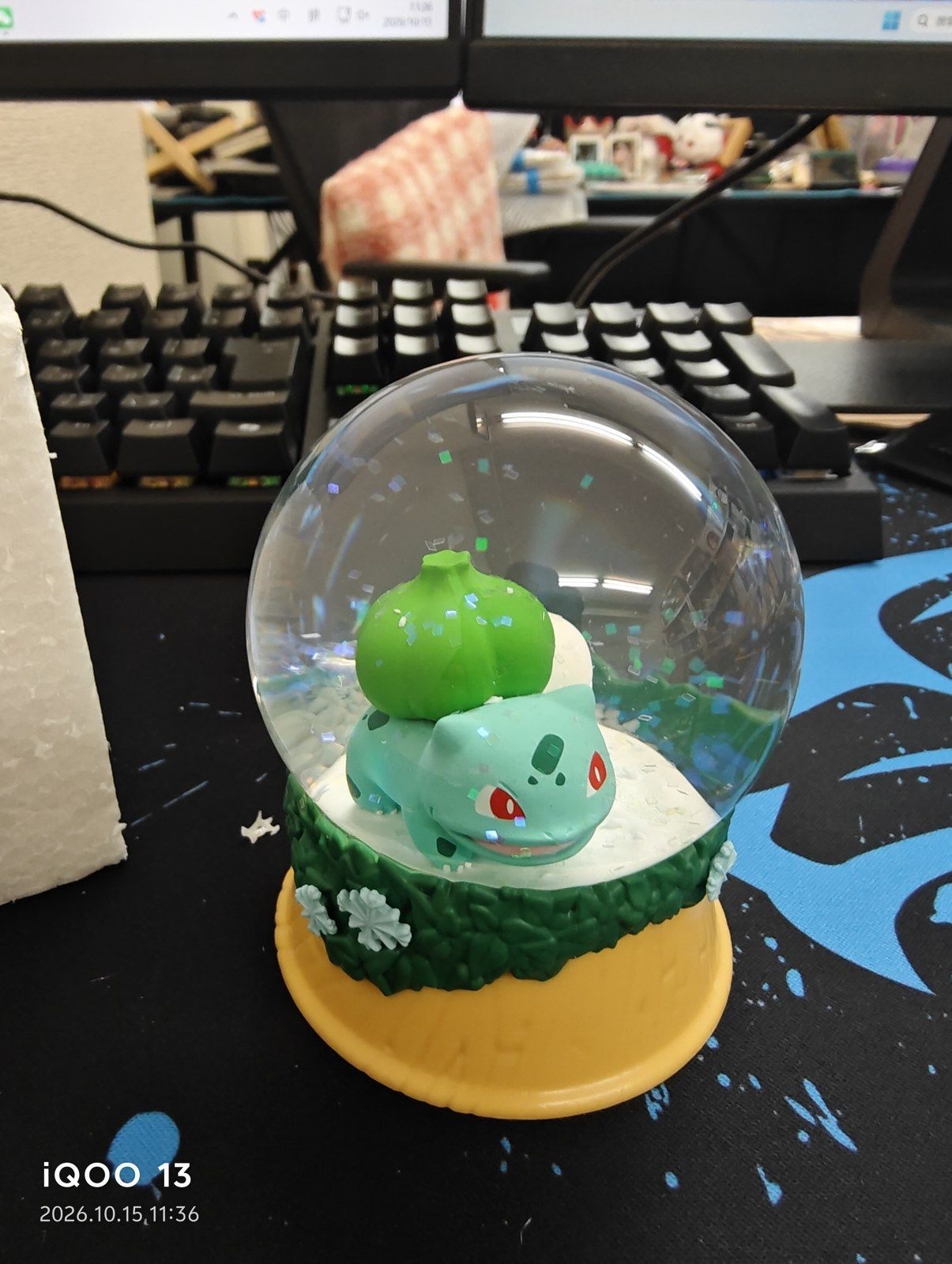 A snow globe with Bulbasaur sitting on white snow inside the clear dome, on a green and yellow base (unedited photo; the camera's date stamp is visible at bottom left)