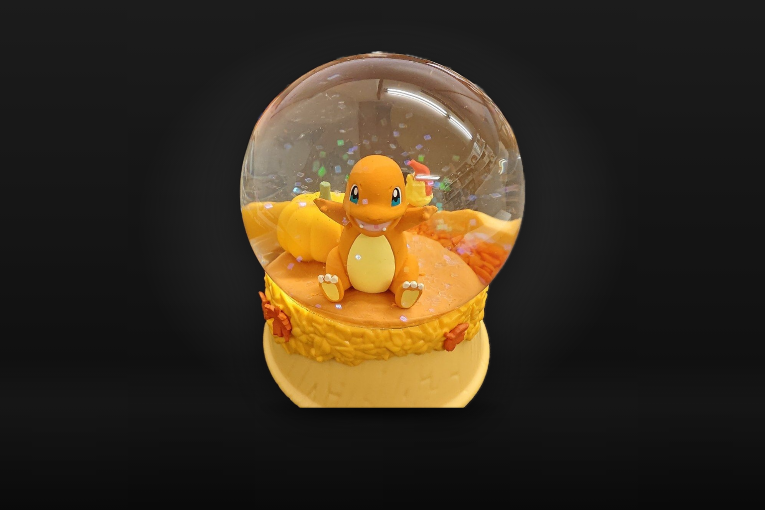 A snow globe on a dark grey background: Charmander sits among an orange pumpkin and autumn leaves inside the clear dome, with holographic glitter, on a yellow base