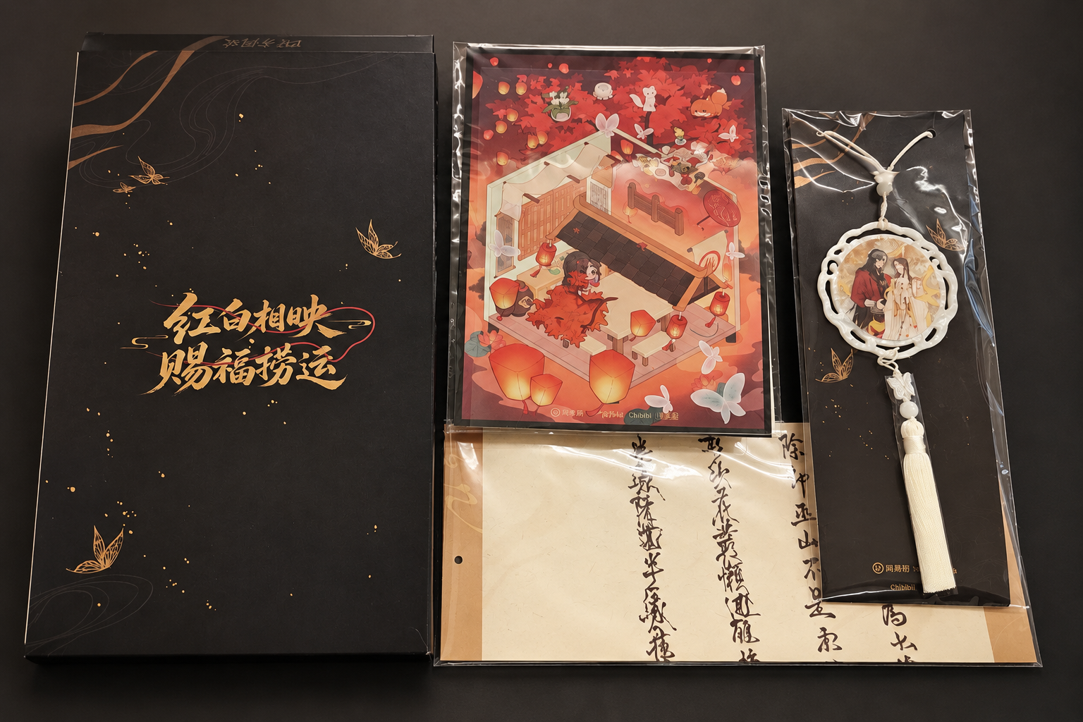 On a dark grey background: the black outer box with gold calligraphy, a red-leaf illustrated sticker, a long letter with calligraphy, and a round acrylic charm of two characters with a white tassel, each in clear bags