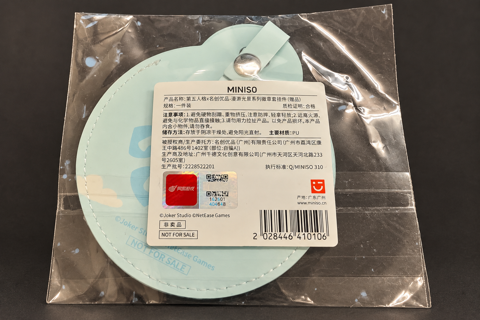 On a dark grey background: the back of the round mint-green badge cover in a clear bag, with a snap-button strap and a white MINISO label printed in Chinese with a NetEase Games sticker