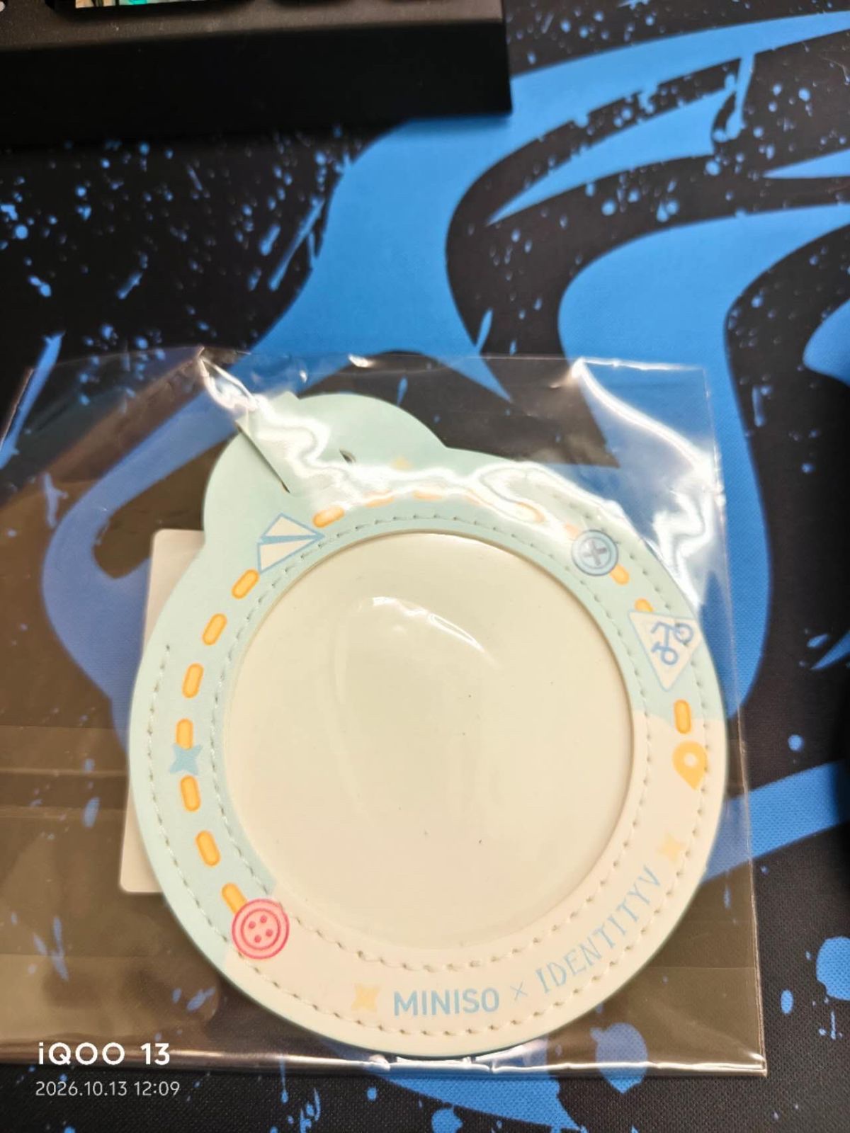 黒と青の敷物の上に置かれた、透明な袋入りの丸い缶バッジカバー。ミントグリーンとアイボリーの縁にオレンジの点線、紙飛行機やボタンの絵、「MINISO × IDENTITYV」の文字がある（未加工の写真、左下にカメラの撮影日時表示あり）