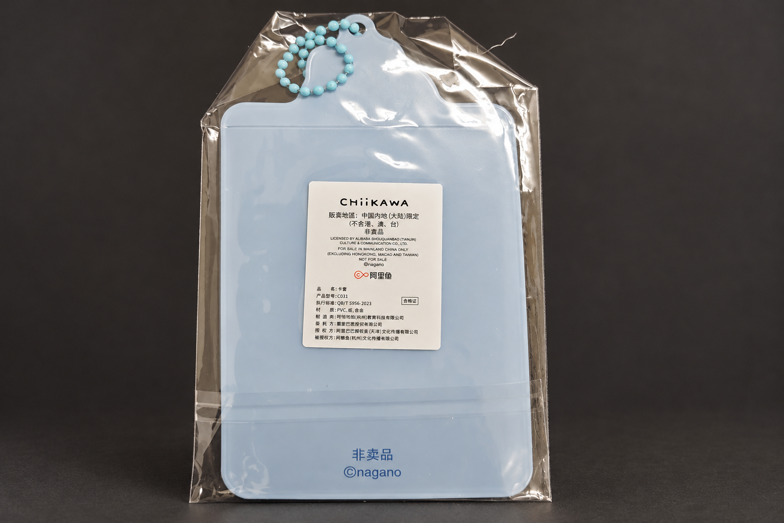 The back of a light-blue card case in a clear bag against a dark grey background, with a light-blue ball chain and a white label printed in Chinese and English, plus the words 非卖品 and ©nagano