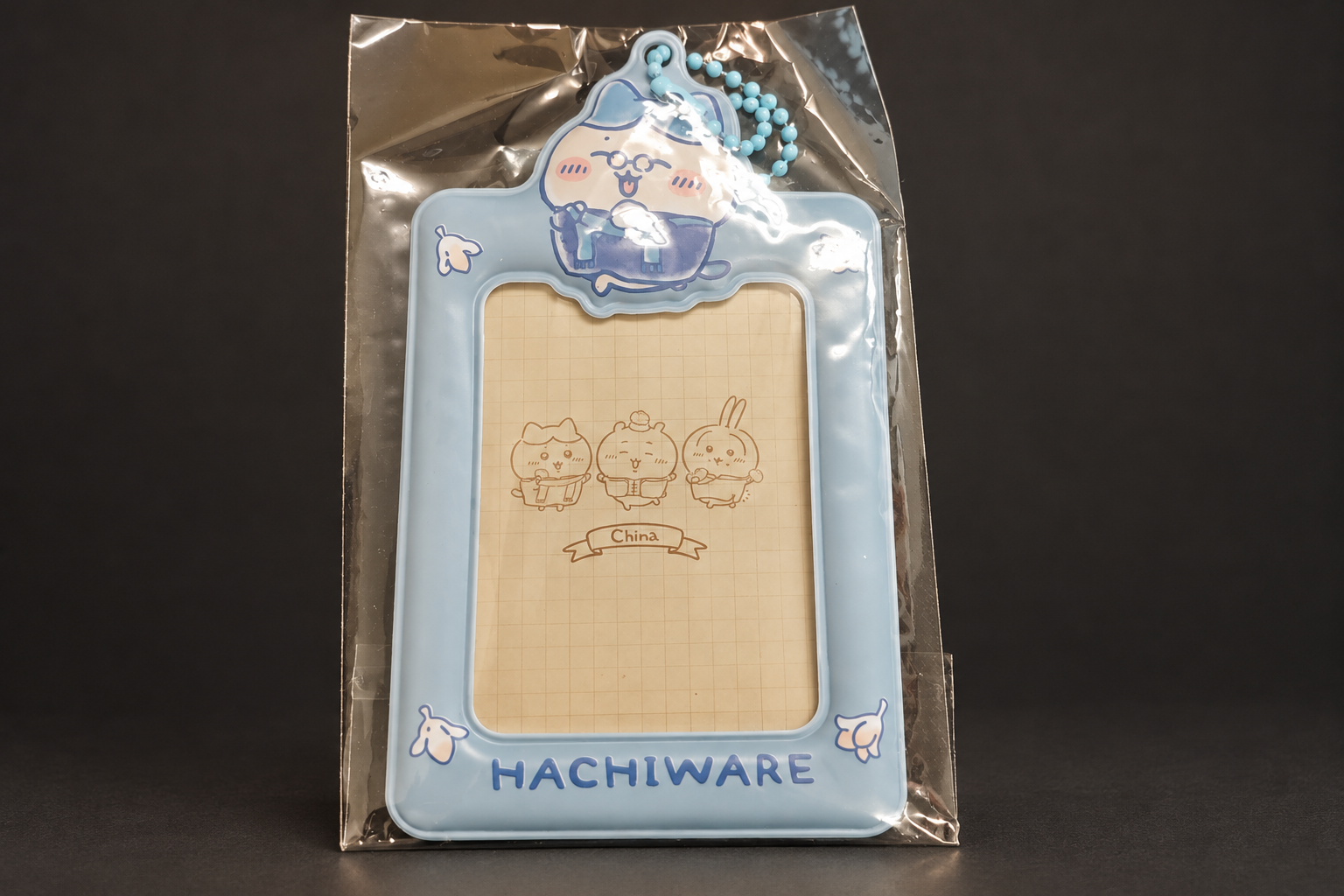 暗いグレーの背景に置かれた、透明な袋入りの水色のカードケース。上にハチワレのダイカットパーツと水色のボールチェーン、下に「HACHIWARE」の文字があり、窓の中に「China」と書かれた方眼の台紙が見える