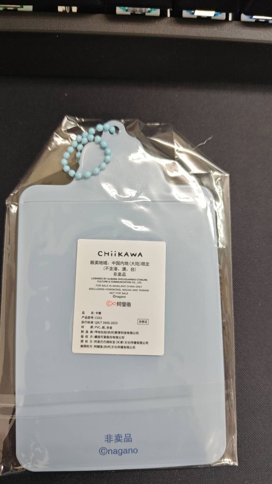 透明な袋に入ったカードケースの裏側。水色の本体に白いラベルが貼られ、「CHiIKAWA」のロゴ、中国語と英語の販売地域や非売品の表示、品名や型番などが書かれている。下部に「非卖品」「©nagano」の印刷がある(未加工の写真、上部にキーボードが写っている)