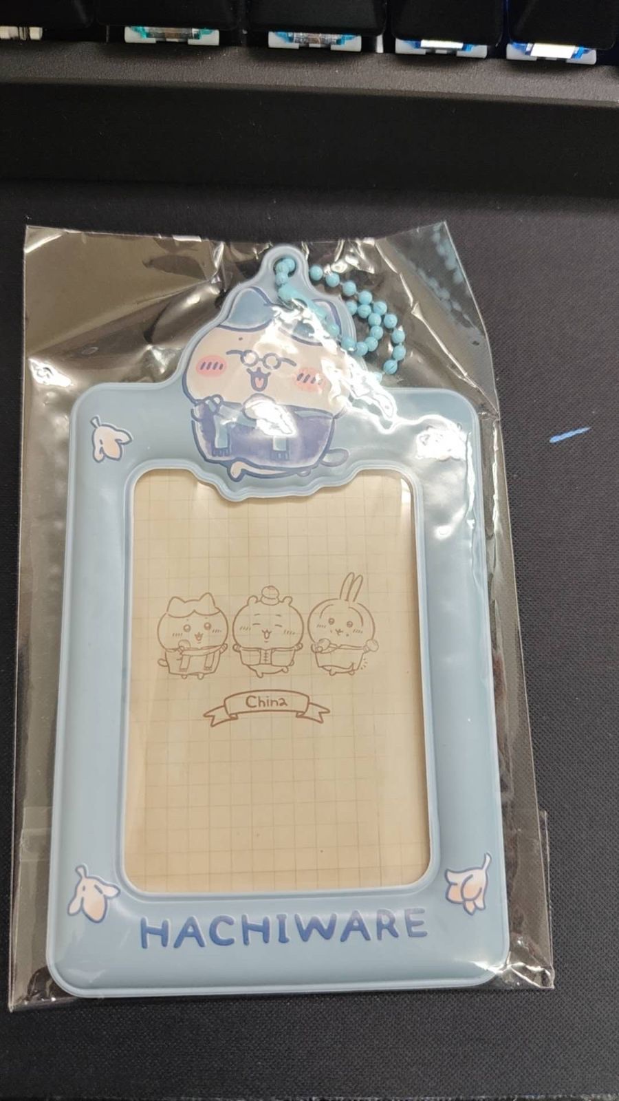 黒いマットの上に置かれた、透明な袋入りの水色のカードケース。上にメガネをかけたハチワレのダイカットパーツと水色のボールチェーン、窓の中にちいかわ・ハチワレ・うさぎと「China」の文字が描かれた方眼の台紙、下に「HACHIWARE」の文字がある(未加工の写真、上部にキーボードが写っている)