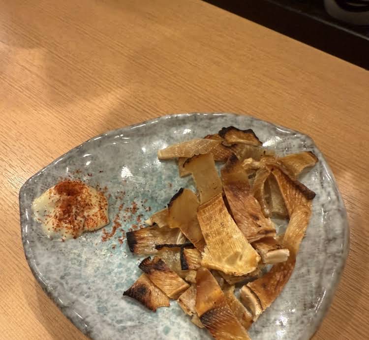 Grilled strips of dried ray fin and mayonnaise with shichimi on a blue-grey plate on a wooden table (unedited photo)