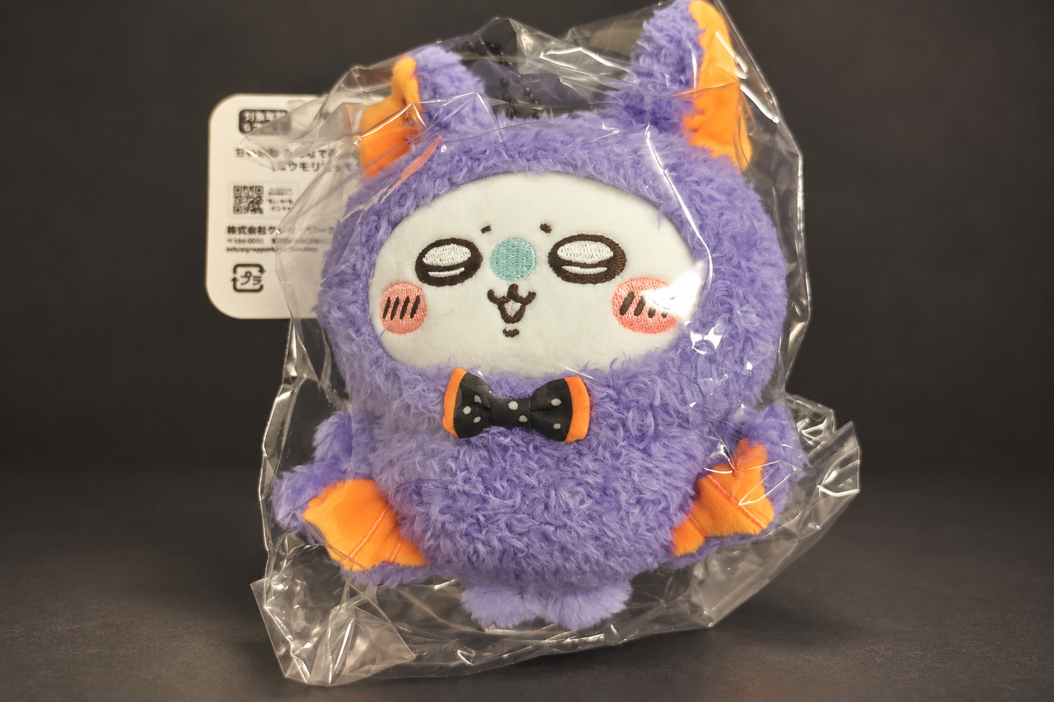 A Momonga mascot in a clear bag against a dark grey background, dressed in a fluffy purple bat costume with orange ears and wings and a black polka-dot bow tie, with a white header tag at the top left