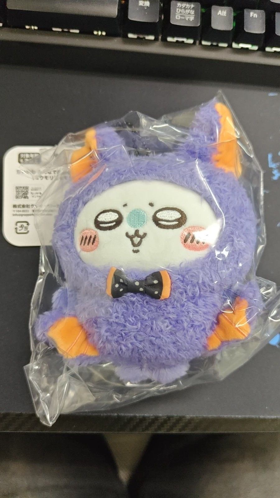Bat Momonga mascot in a clear bag on a black mat, in a fluffy purple bat suit with orange ears and wings and a black polka-dot bow tie, with the header tag at the top left (unedited photo; a keyboard is visible at the top)