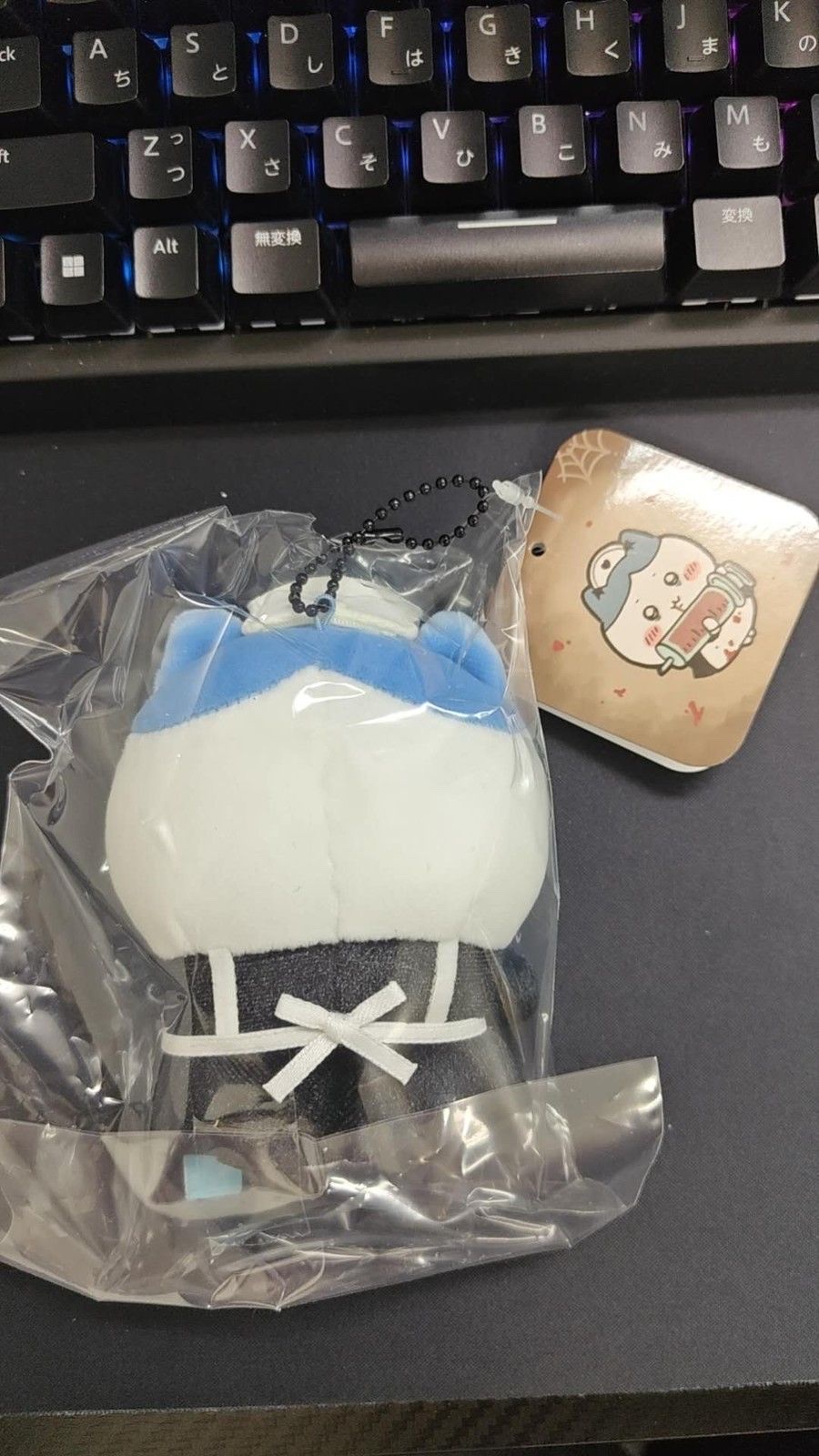 The back of Nurse Hachiware in its clear bag, with the white apron strings tied at the back, the ball chain at the top and the illustrated back of the square tag at the top right (unedited photo; a keyboard is visible at the top)