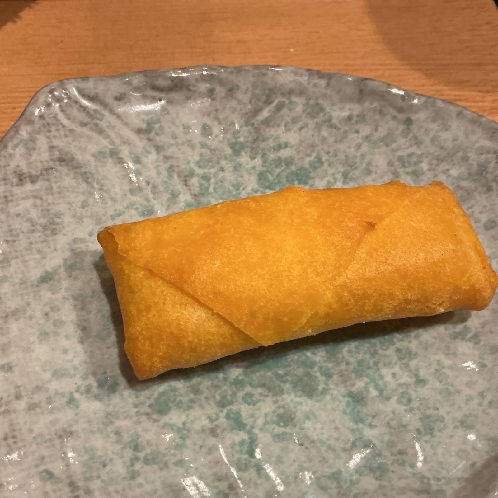 A golden fried spring roll on a grey plate with green speckles, on a wooden table (unedited photo)