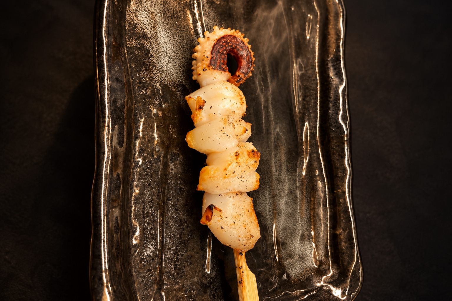 A grilled squid skewer on a long black-glazed plate against a dark background, with the curled tentacle section at the top