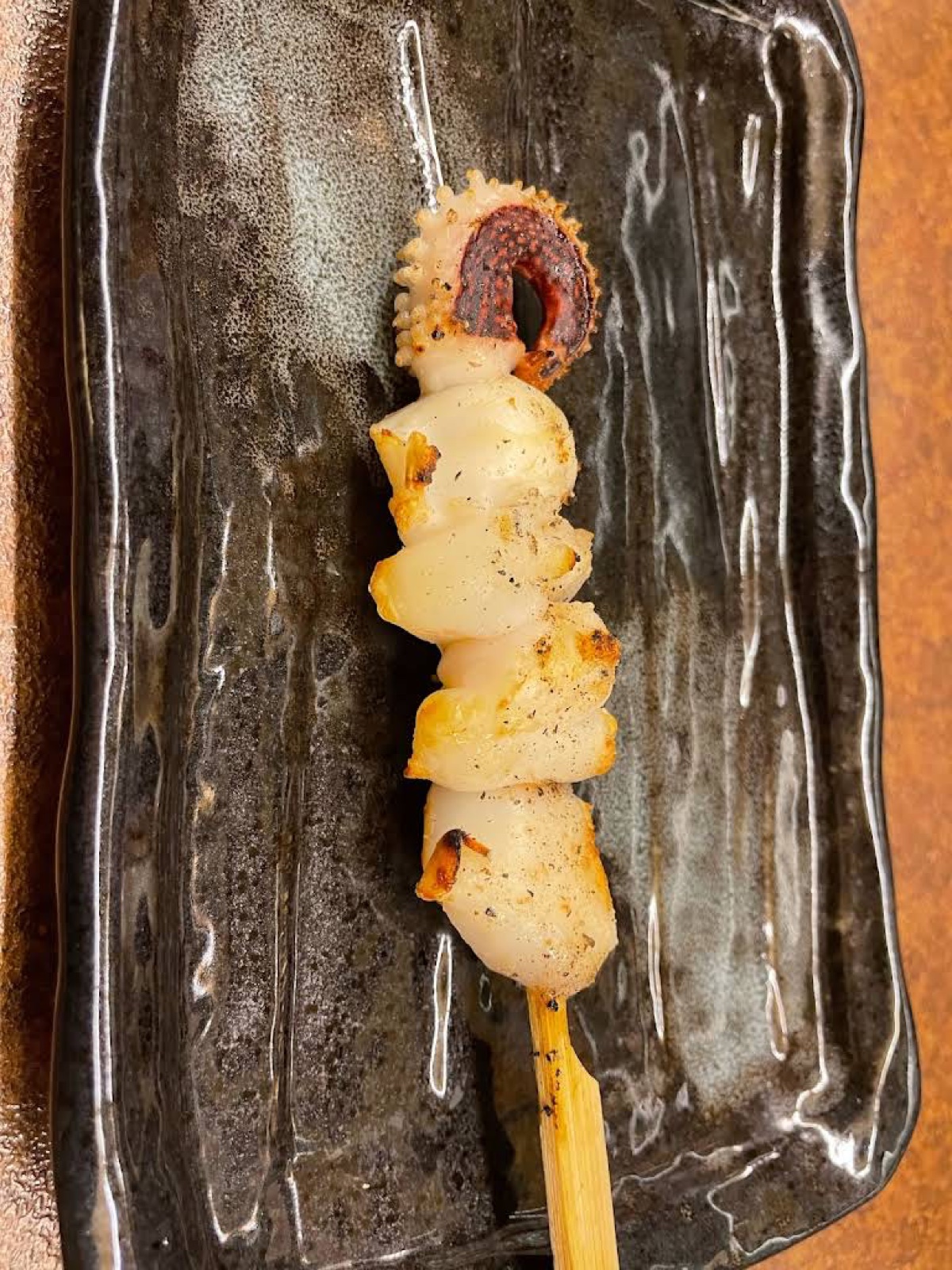 木のテーブルに置かれた黒い釉薬の長皿に、焼き目のついたいかの串が1本のっている（未加工の写真）