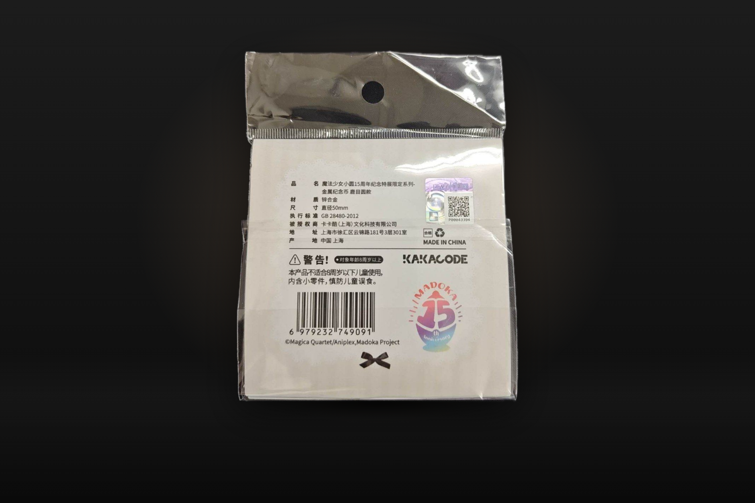 Against a dark backdrop, the back of the coin's packaging: a white card with a Chinese product label, a holographic sticker with a QR code, a barcode and the MADOKA 15th anniversary logo