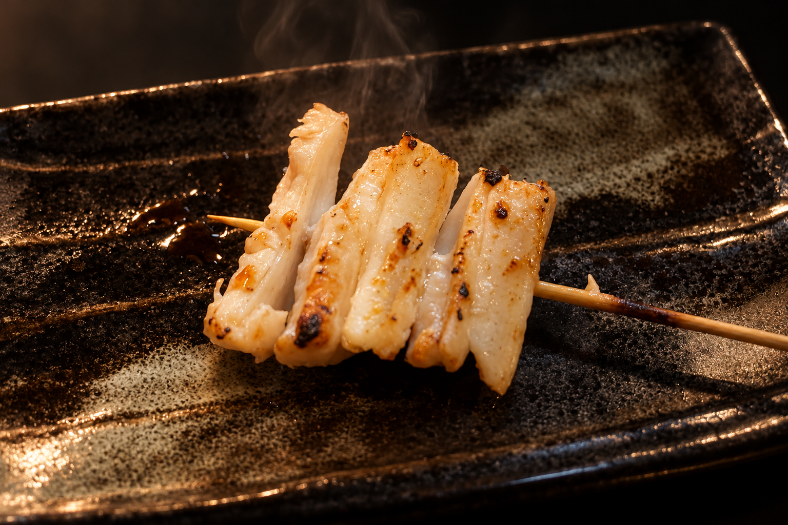 A single skewer of grilled white leek with charred spots on a long black-glazed plate against a dark background