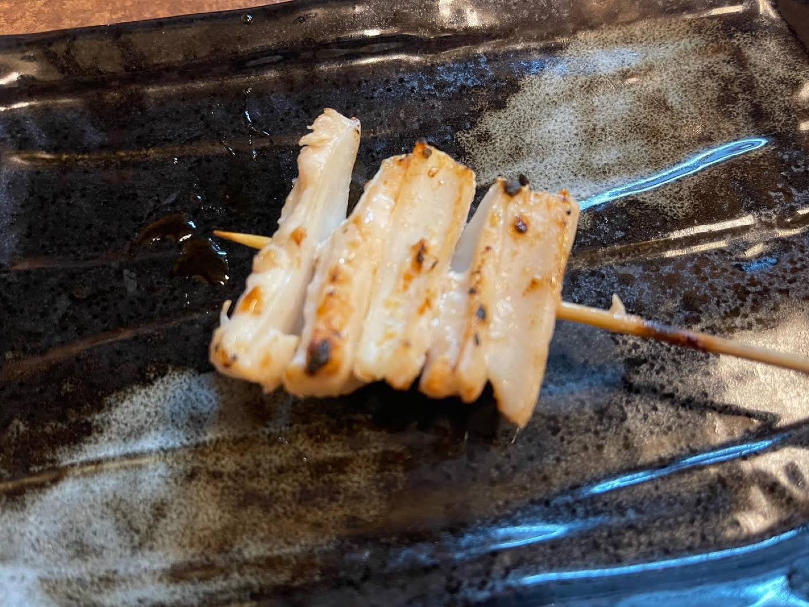 黒い長皿の上に、焼き色のついた白ねぎの串が1本置かれている。切り口に何層も重なった層が見える