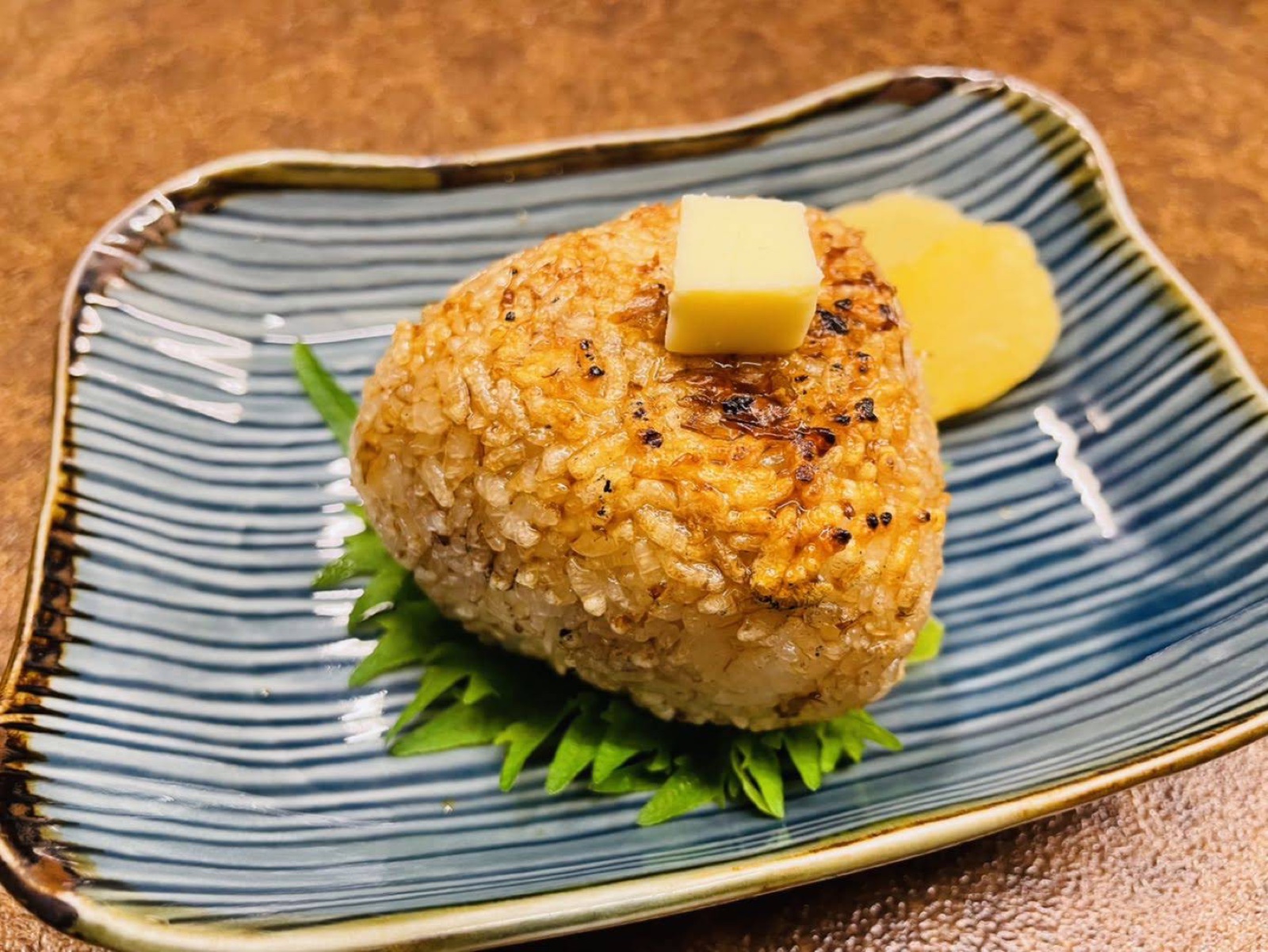 A browned triangular grilled rice ball on a blue striped dish, with a cube of butter on top, pickled radish beside it and green leaves underneath