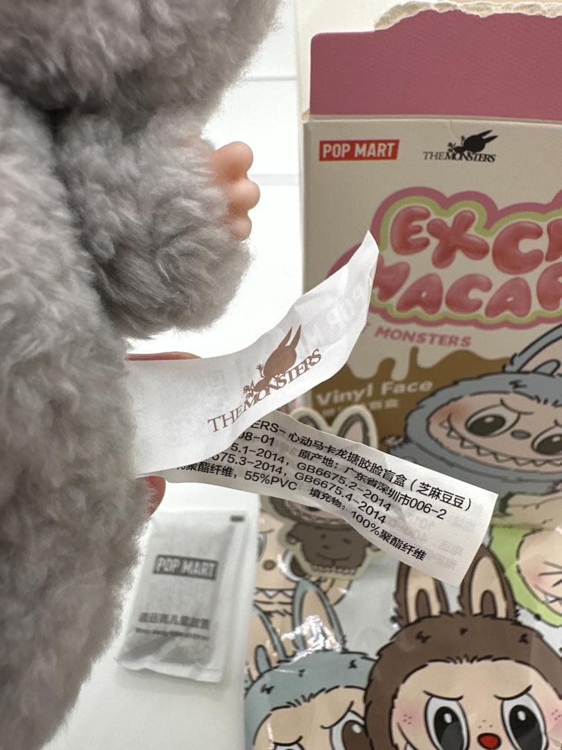 Close-up of a grey LABUBU plush body and fabric labels beside its opened box