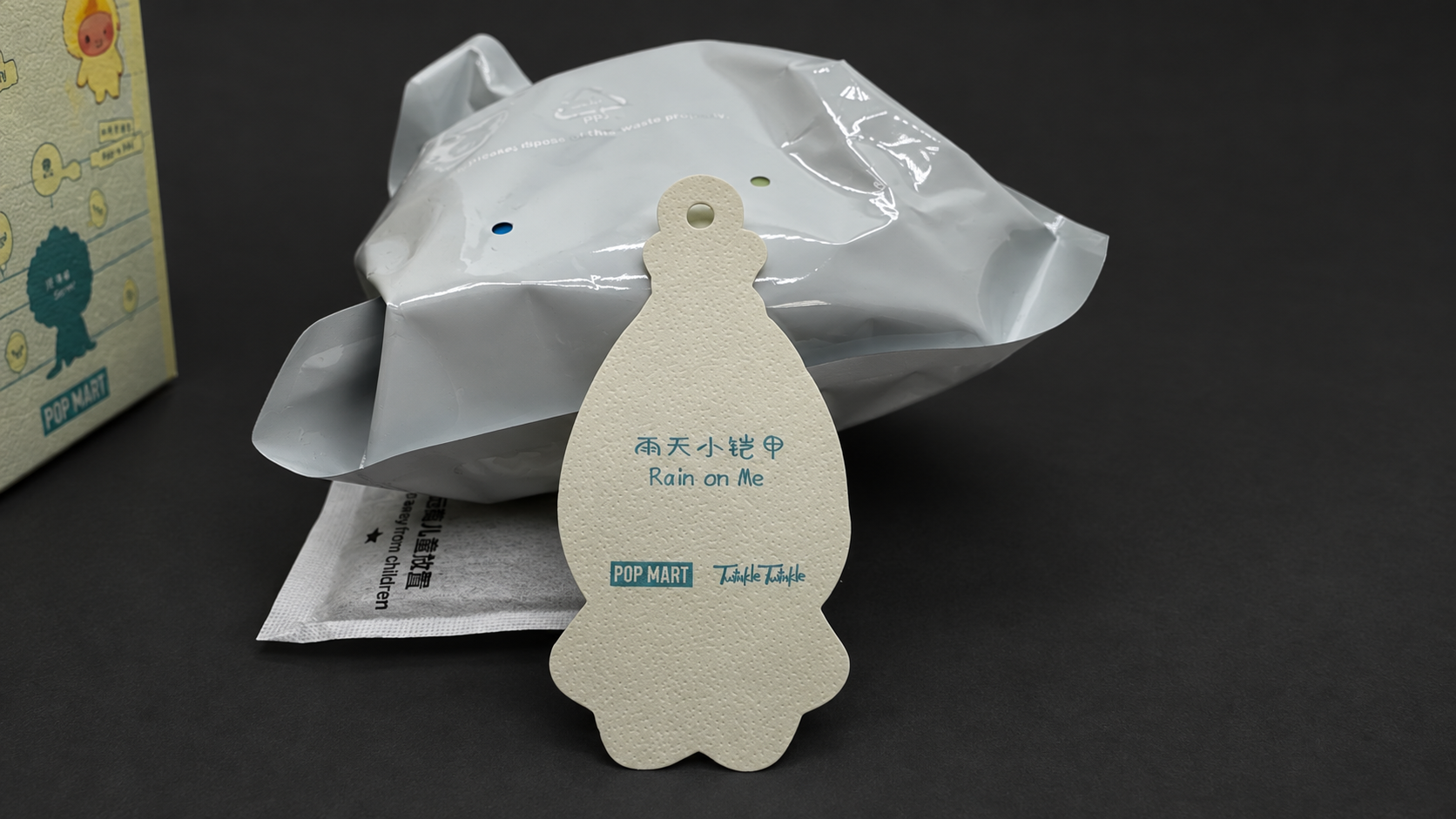 Background-edited photo of the reverse of a Rain on Me character card beside a silver inner bag