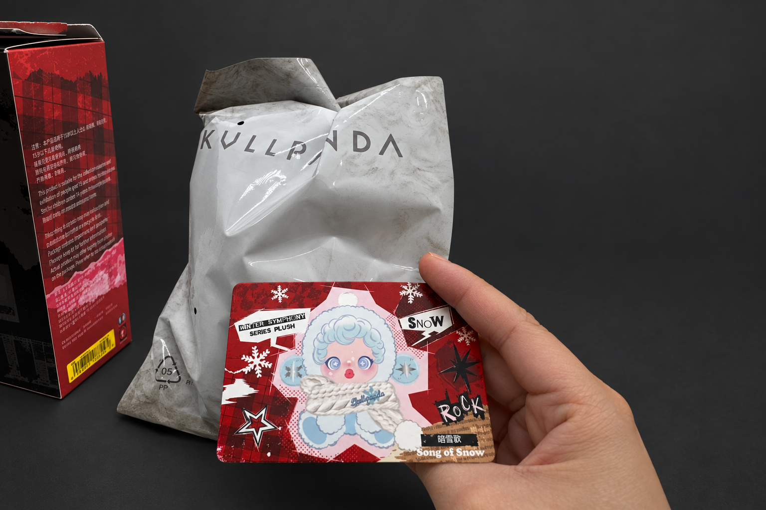 Edited photo of a hand holding a Song of Snow card beside the bag and box